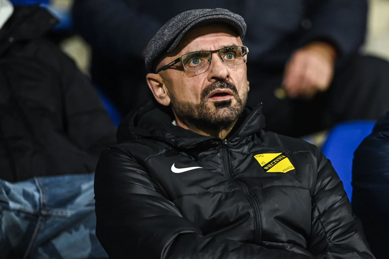 Mężczyzna w okularach i kaszkiecie, ubrany w sportową kurtkę z żółtym logotypem sportowego klubu, siedzi na trybunach i patrzy uważnie przed siebie, w tle widoczne są inne osoby i niebieskie fotele stadionowe.