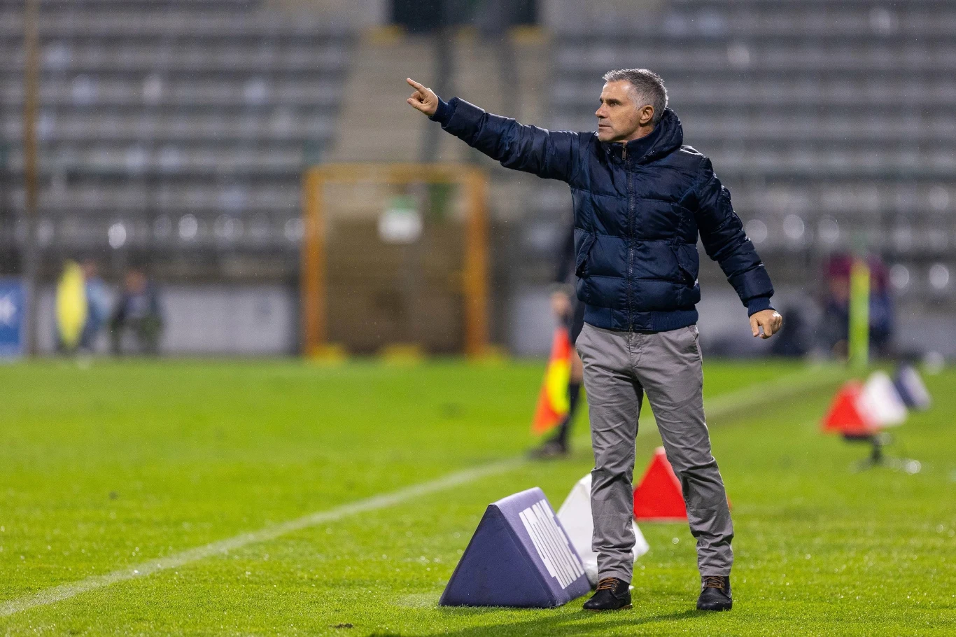 Mężczyzna w sportowej kurtce i spodniach stoi przy linii bocznej boiska piłkarskiego, energicznie gestykulując w stronę pola gry, w tle widoczne są trybuny oraz znaczniki boiska.