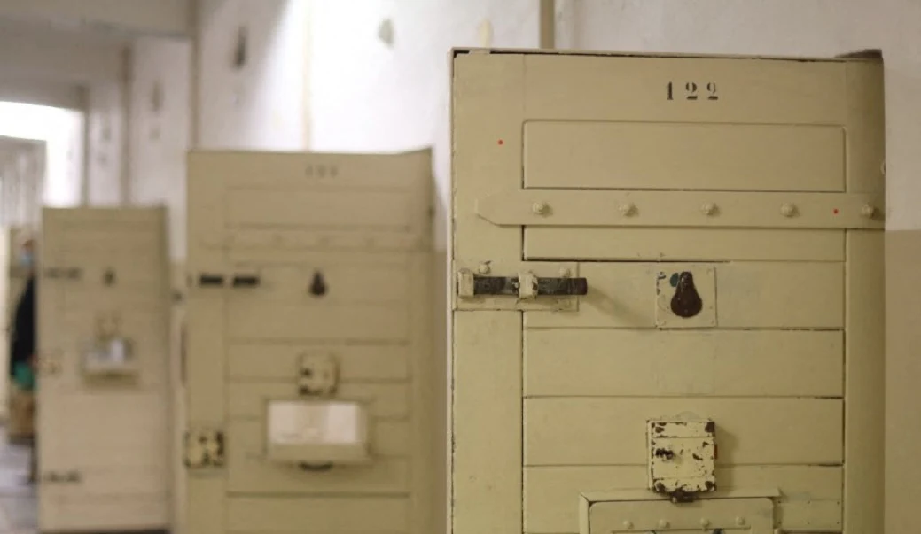 Ciężkie, metalowe drzwi do cel ustawione w szeregu w jasnym, wąskim korytarzu więziennym, każdy z licznymi zamkami oraz niewielkim wizjerem.