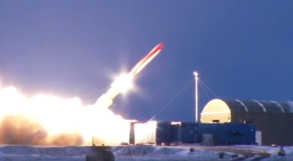 Startująca rakieta wynurza się z wyrzutni, generując jasną smugę płomienia i dymu na tle nieba, obok widoczne są zabudowania infrastruktury technicznej podświetlone nocnym oświetleniem.