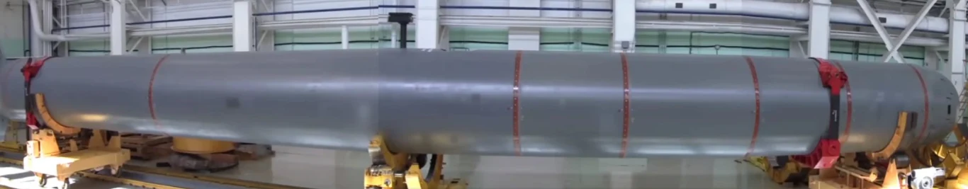 Torpeda o napędzie nuklearnym "Posejdon". Nie są publicznie znane jej dokładne wymiary, ale jest długa nawet na 20 metrów Długi, cylindryczny obiekt techniczny o szarej powierzchni i metalicznych elementach, umieszczony na żółtych podporach w jasnej hali przemysłowej.