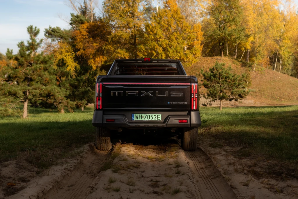 Czarny pickup Maxus z zieloną tablicą rejestracyjną stoi na piaszczystej drodze otoczonej trawą, drzewami iglastymi oraz liściastymi w intensywnych, jesiennych barwach. Tylne światła są wyeksponowane, a pojazd skierowany jest tyłem do obiektywu.
