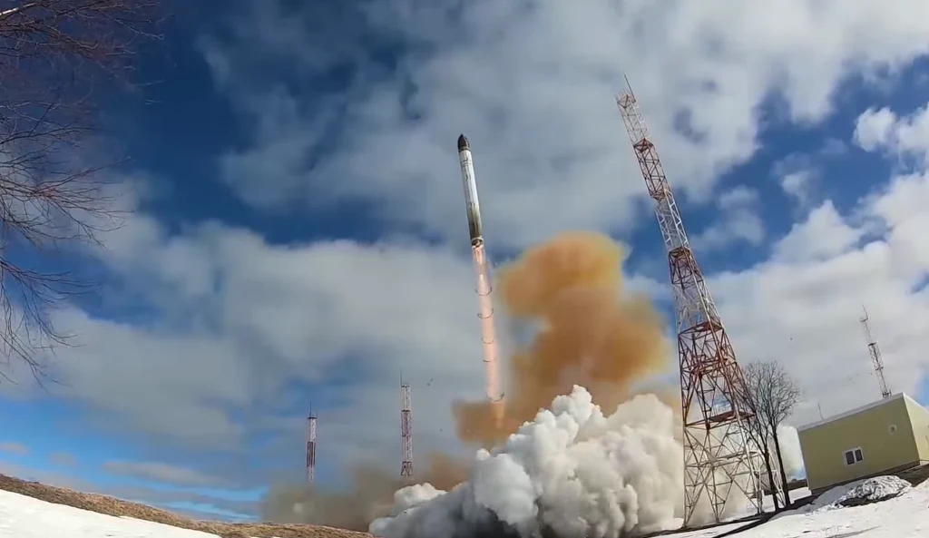 Rakieta w momencie startu otoczona chmurą dymu, unosząca się pionowo w górę, w tle wieże komunikacyjne oraz budynek, niebo zachmurzone z przebłyskami błękitu, na ziemi pozostałości śniegu.