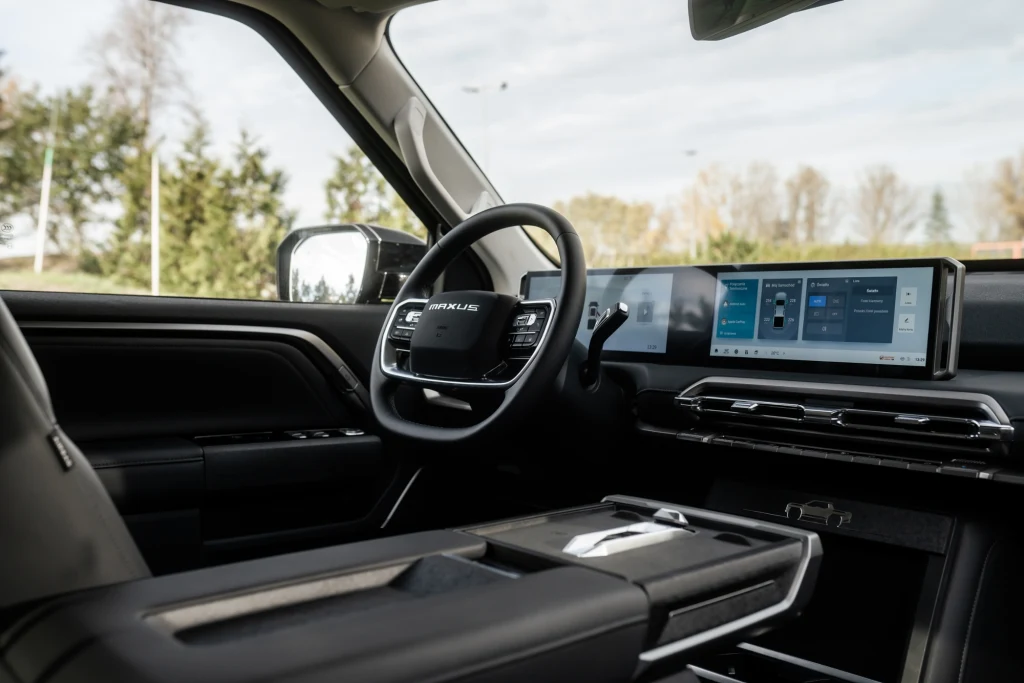 Luksusowe wnętrze samochodu z nowoczesną deską rozdzielczą, dużym dotykowym ekranem multimedialnym oraz kierownicą z logo marki Lexus. W tle widoczne delikatnie rozmyte drzewa za szybą.