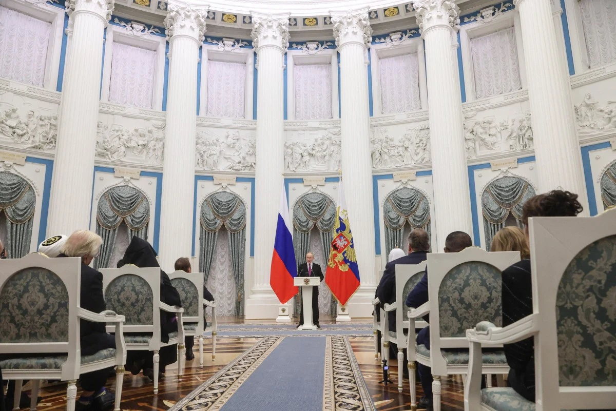 Władimir Putin podczas wystąpienia na Kremlu Oficjalna ceremonia w okazałej sali z wysokimi białymi kolumnami, gdzie naprzeciw zgromadzonych osób stoi mężczyzna przy mównicy na tle dwóch flag, rosyjskiej i regionalnej, całość utrzymana w wystawnej, klasycznej stylistyce.