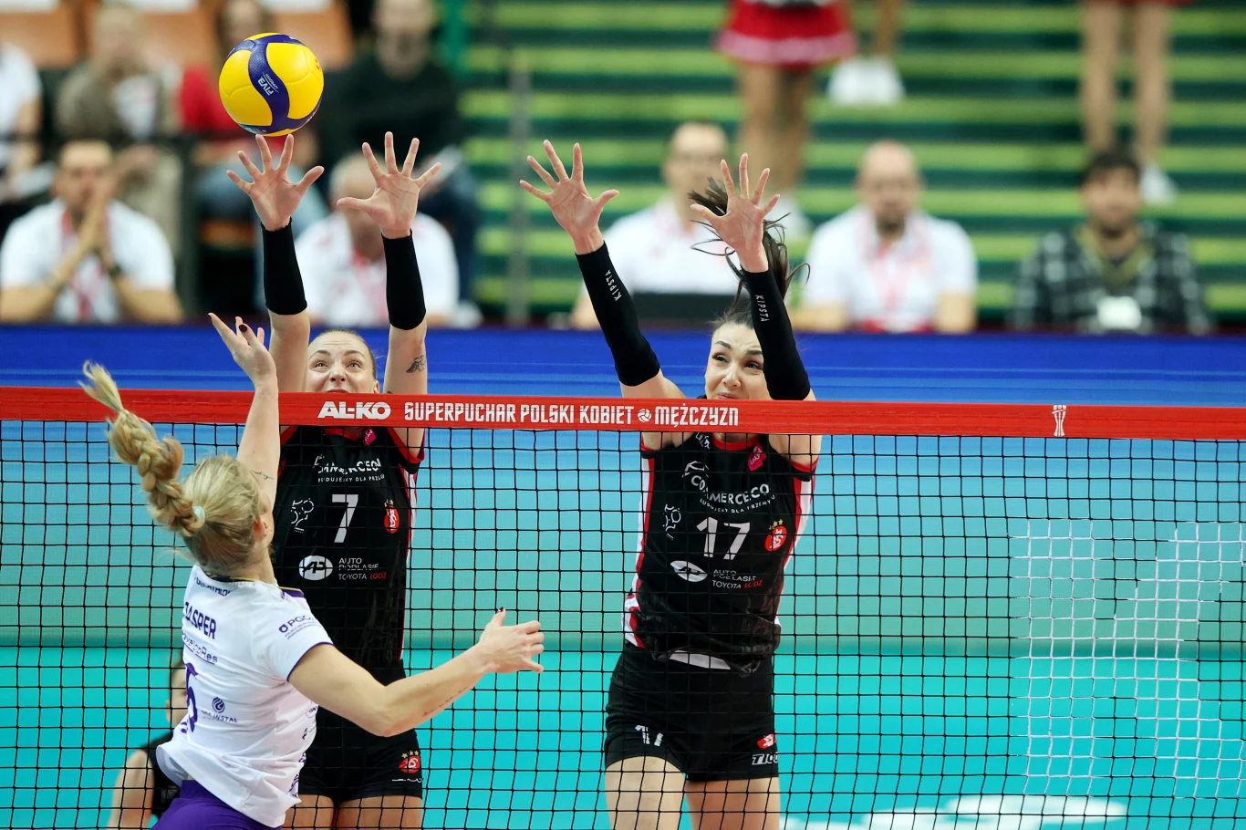 Dwie siatkarki w czarnych strojach próbują zablokować atak zawodniczki drużyny przeciwnej podczas meczu siatkówki, po obu stronach siatki widoczne skupienie i dynamika akcji, w tle widoczna publiczność.