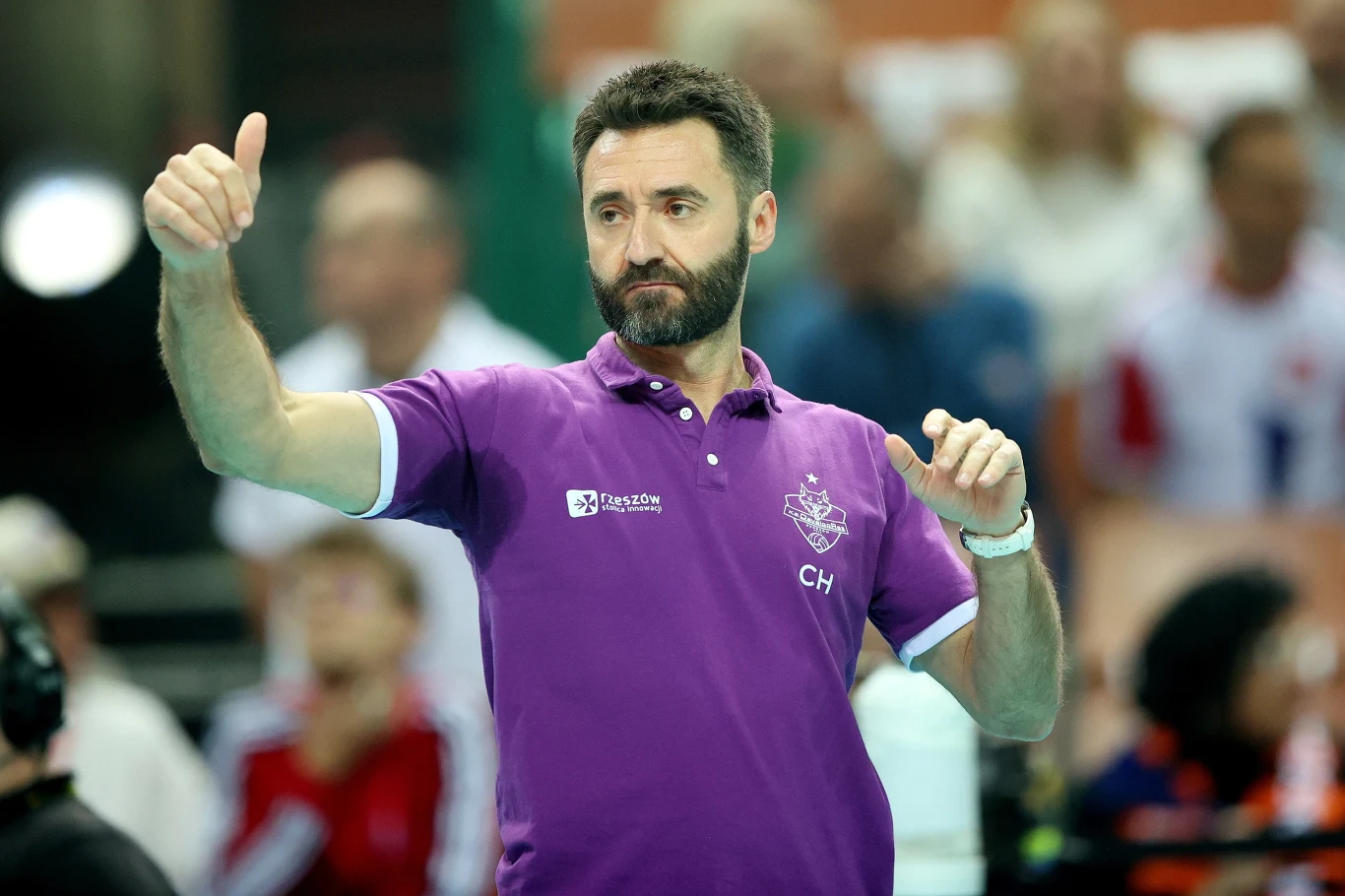 Mężczyzna z brodą ubrany w fioletową koszulkę polo wykonuje gest uniesionego kciuka, na tle rozmytego tłumu osób. Na koszulce widoczne są logo i inicjały, sugerujące przynależność do drużyny sportowej lub organizacji.