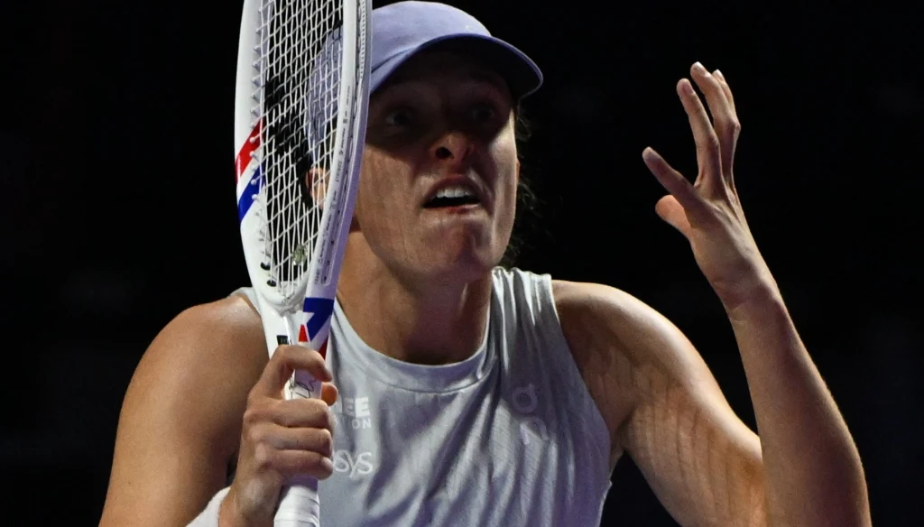 Tenisistka w fioletowej czapce i jasnym stroju sportowym trzyma rakietę tenisową, wyrażając emocjonalną reakcję gestem ręki, z wyraźnym napięciem na twarzy.