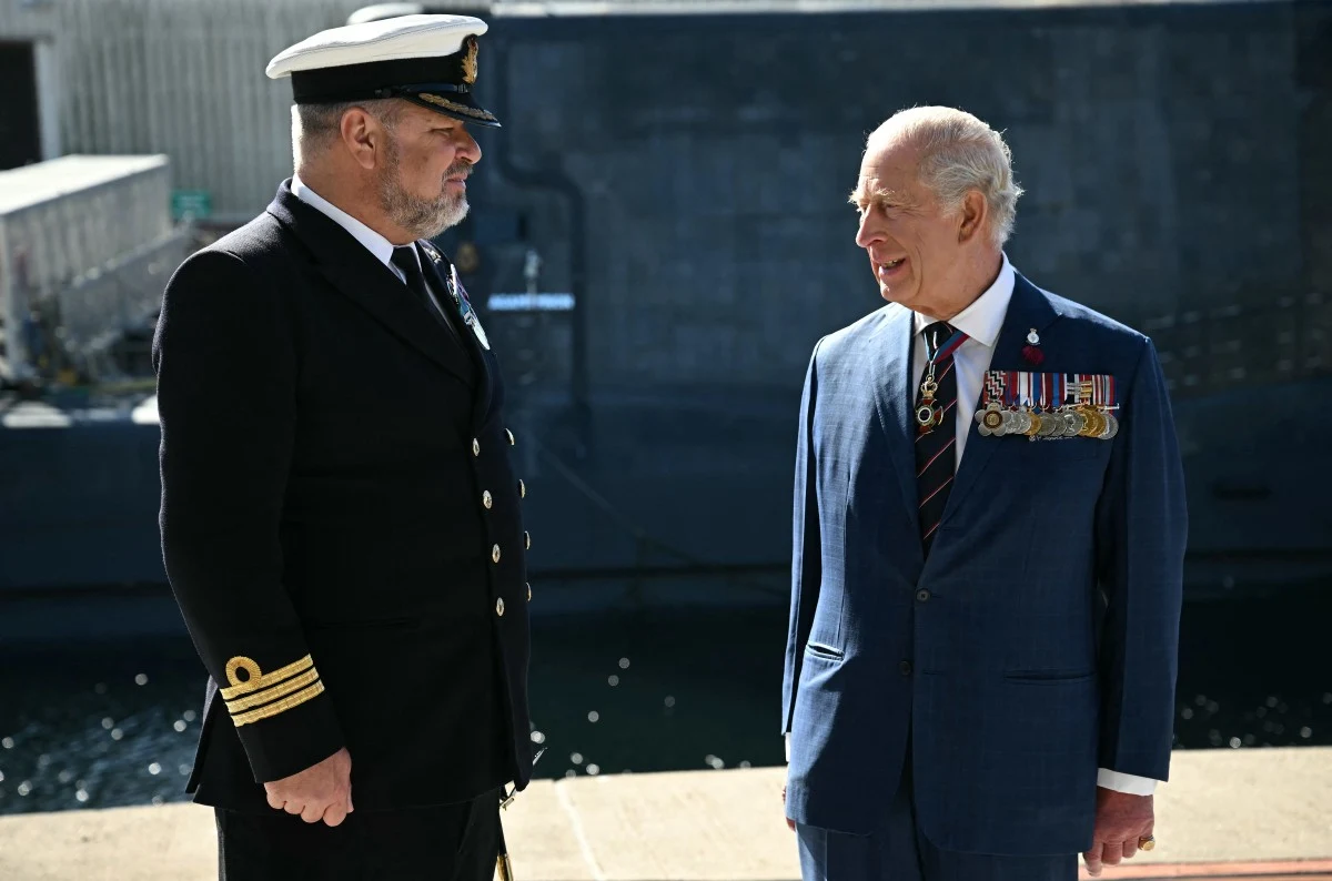 Dwóch mężczyzn w mundurach rozmawia ze sobą na tle okrętu wojskowego, po prawej stoi mężczyzna w niebieskim garniturze z licznymi medalami, po lewej mężczyzna w mundurze marynarki wojennej.