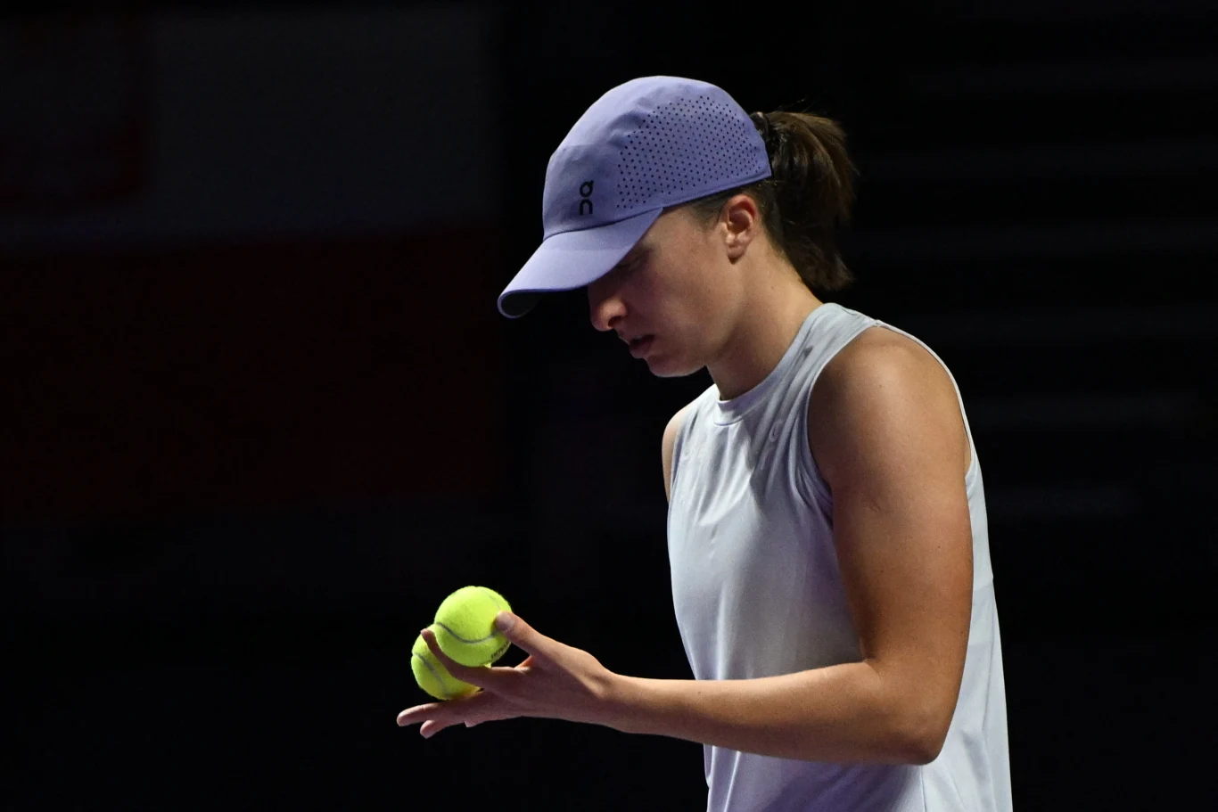 Młoda kobieta w sportowej koszulce i czapce z daszkiem koncentruje się na trzymanych w dłoni piłkach tenisowych, ciemne tło podkreśla postać oraz skupienie.