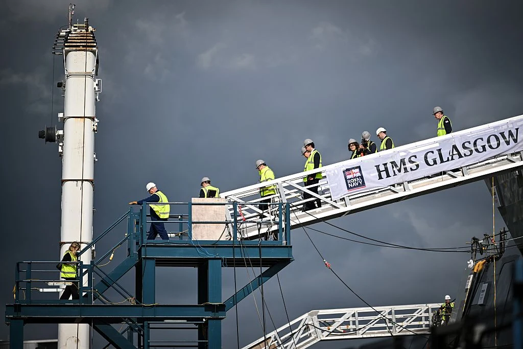 Grupa osób w kaskach ochronnych i kamizelkach odblaskowych wchodzi po rampie z napisem HMS Glasgow na teren statku, w tle ciemne chmury i fragment konstrukcji przemysłowej.