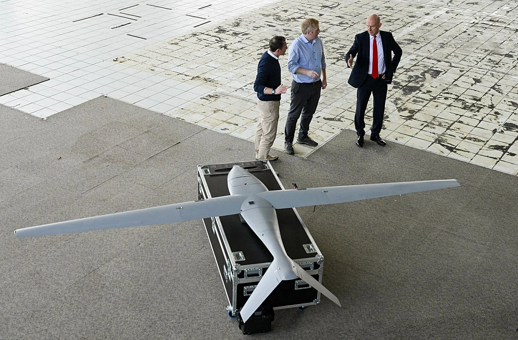 Duży dron leżący na czarnej skrzyni transportowej, obok którego stoją trzej mężczyźni rozmawiający i wskazujący na urządzenie, całość znajduje się na częściowo zniszczonej podłodze z płytek.