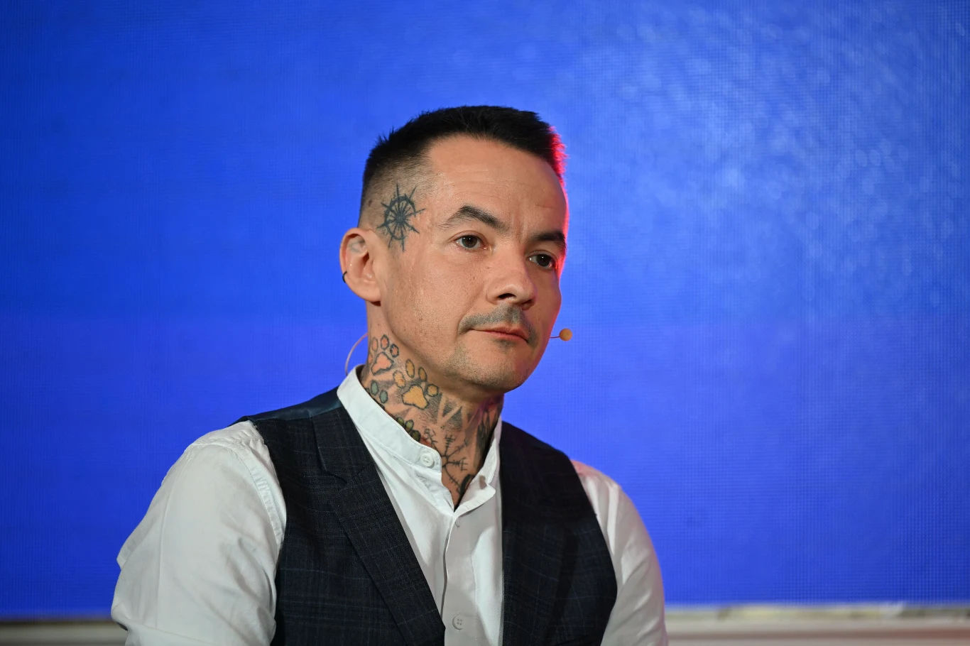 Mężczyzna o ciemnych, krótkich włosach z widocznymi tatuażami na twarzy i szyi, ubrany w białą koszulę i ciemną kamizelkę, siedzi w dobrze oświetlonym wnętrzu na tle niebieskiej ściany, z mikrofonem na twarzy.