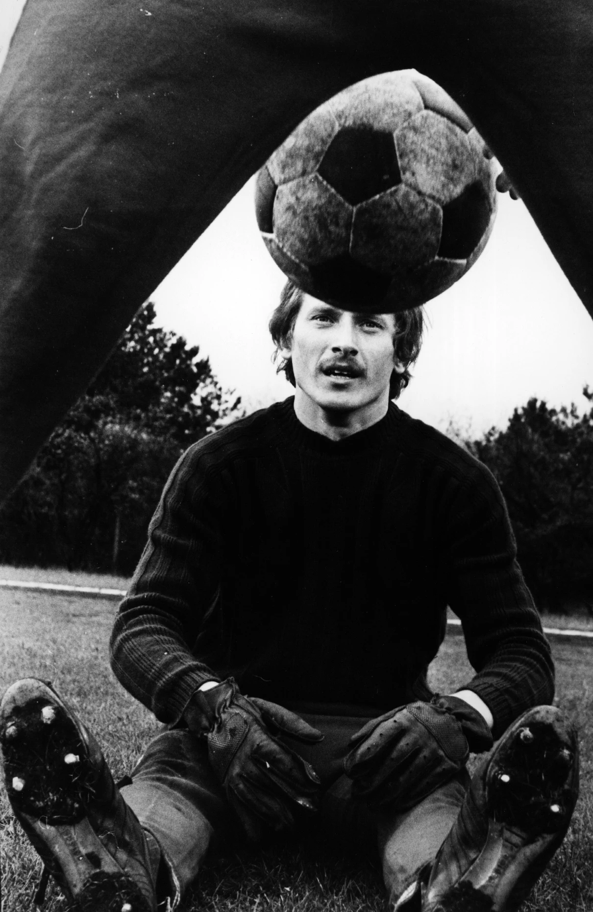 Mężczyzna w stroju bramkarza siedzi na trawie, zakłada rękawice i patrzy w kamerę, nad jego głową zawieszona jest piłka nożna w dynamicznym ujęciu z nogami drugiego piłkarza w kadrze.