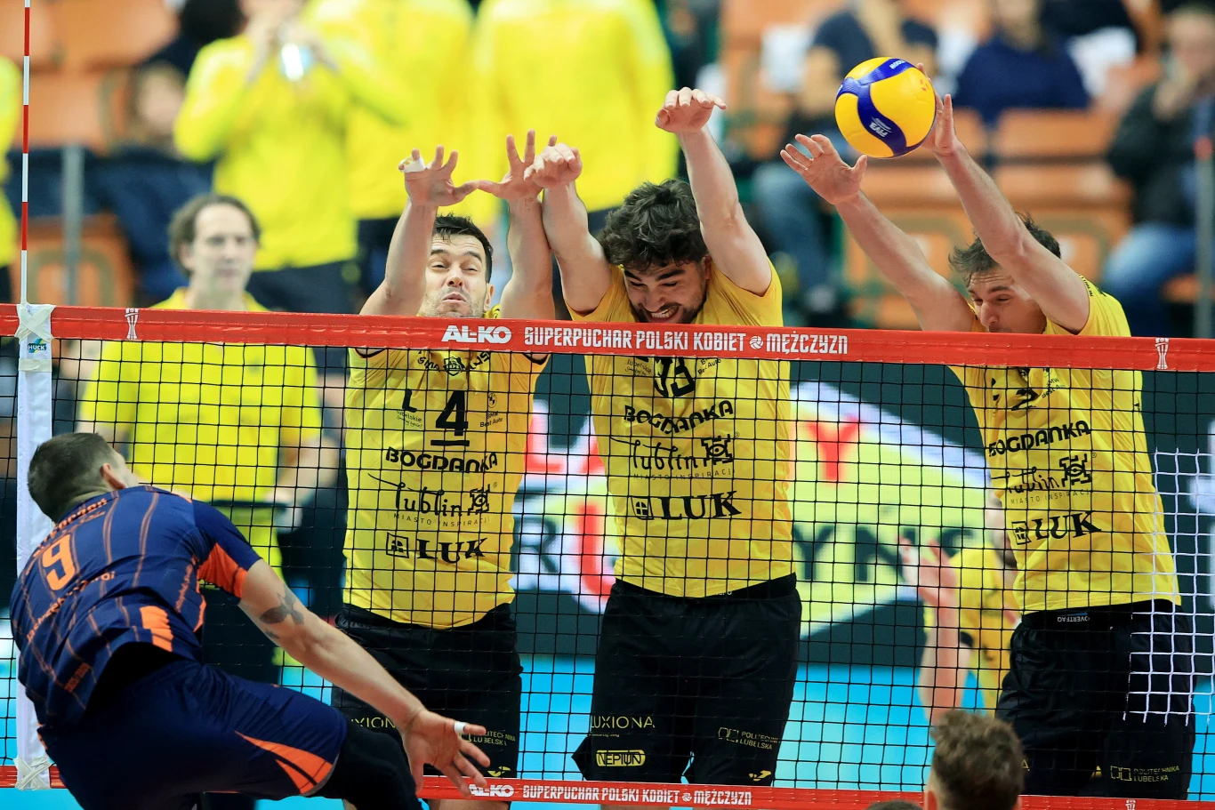 Trzech siatkarzy w żółtych koszulkach wykonuje blok przy siatce podczas akcji meczowej, naprzeciwko zawodnik w niebiesko-pomarańczowej koszulce atakuje piłkę, w tle widoczni kibice i sędziowie.