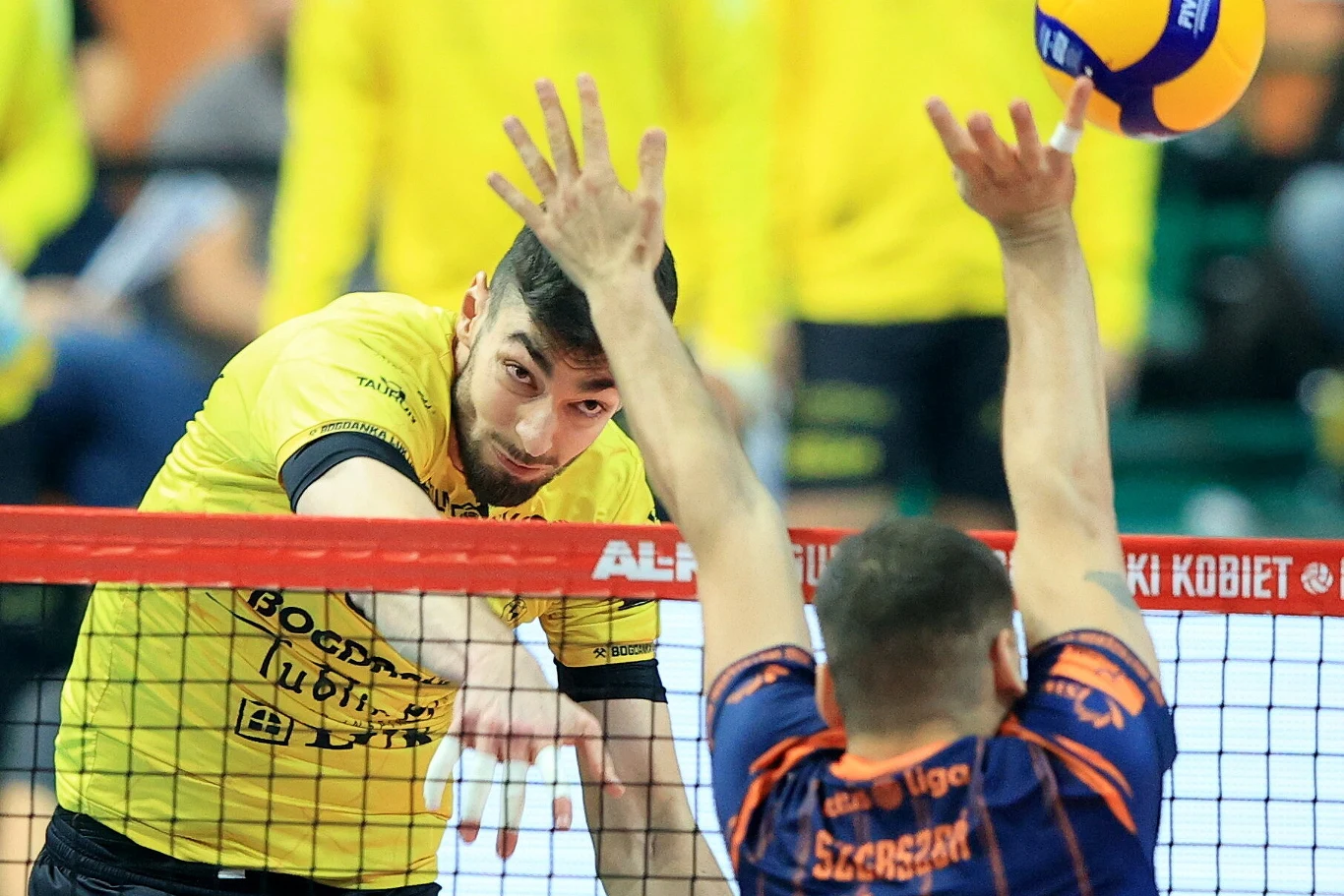 Zawodnik w żółtej koszulce atakuje piłkę przez siatkę, podczas gdy przeciwnik próbuje zablokować piłkę unosząc ręce na wysokość siatki. W tle pozostali uczestnicy meczu, dominują kolory żółty i niebieski.