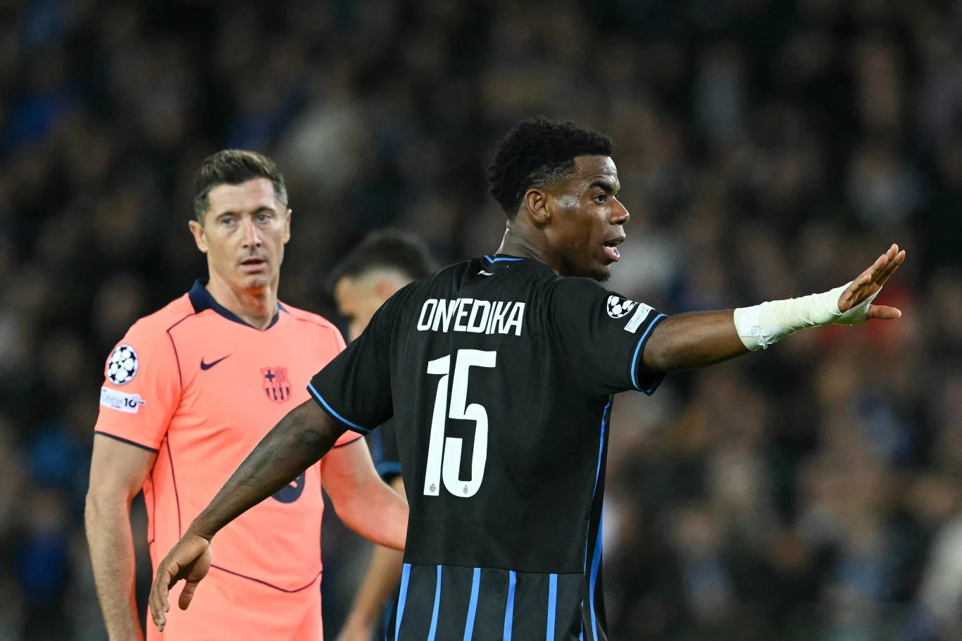 Dwóch piłkarzy podczas meczu piłki nożnej, jeden z nich ubrany w czarny strój z numerem 15 wykonuje gest ręką, drugi w różowej koszulce stoi w tle.