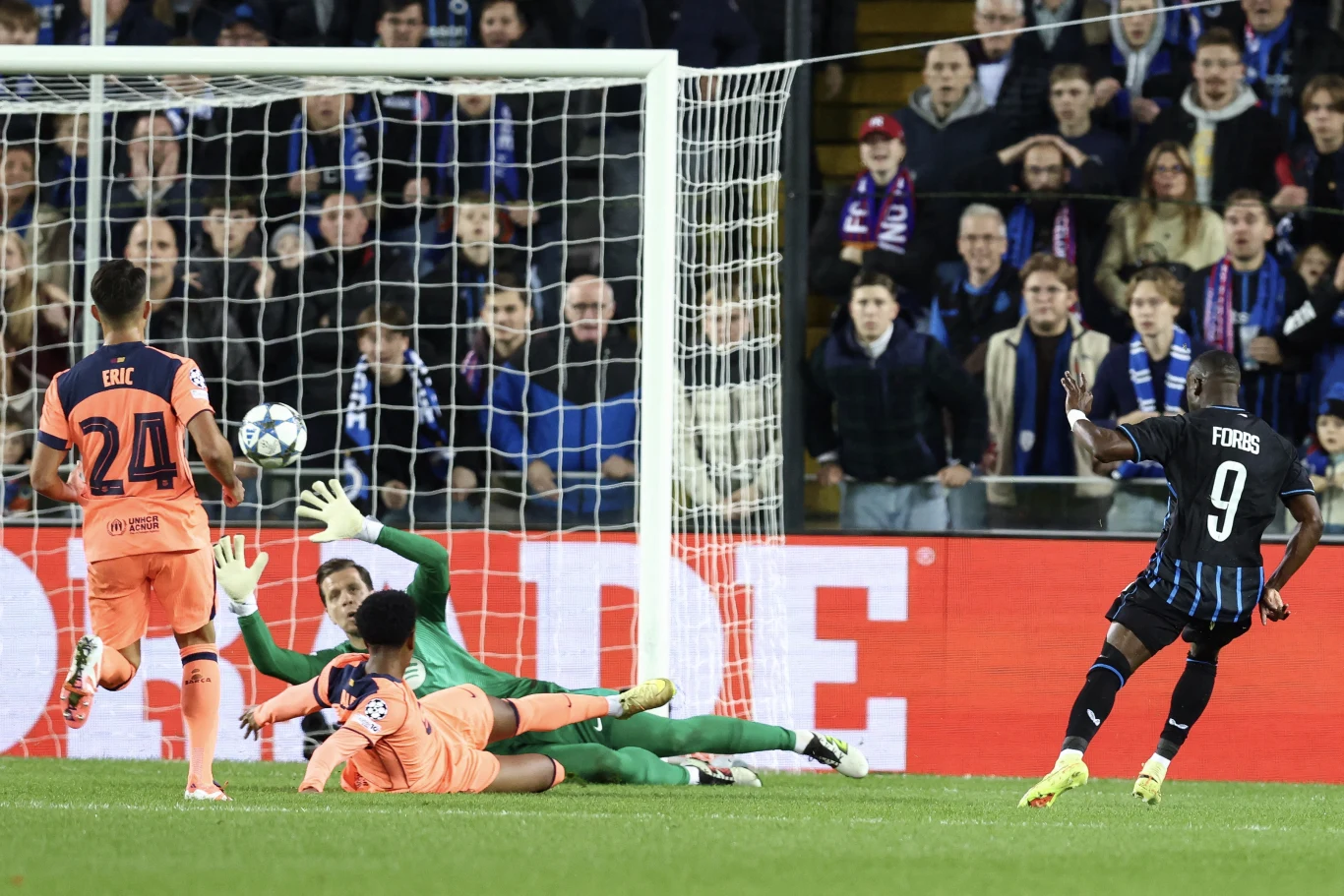 Piłkarz w czarnej koszulce strzela na bramkę, bramkarz i jeden z obrońców w pomarańczowych strojach próbują zablokować piłkę, a w tle widoczna jest grupa kibiców na trybunach.