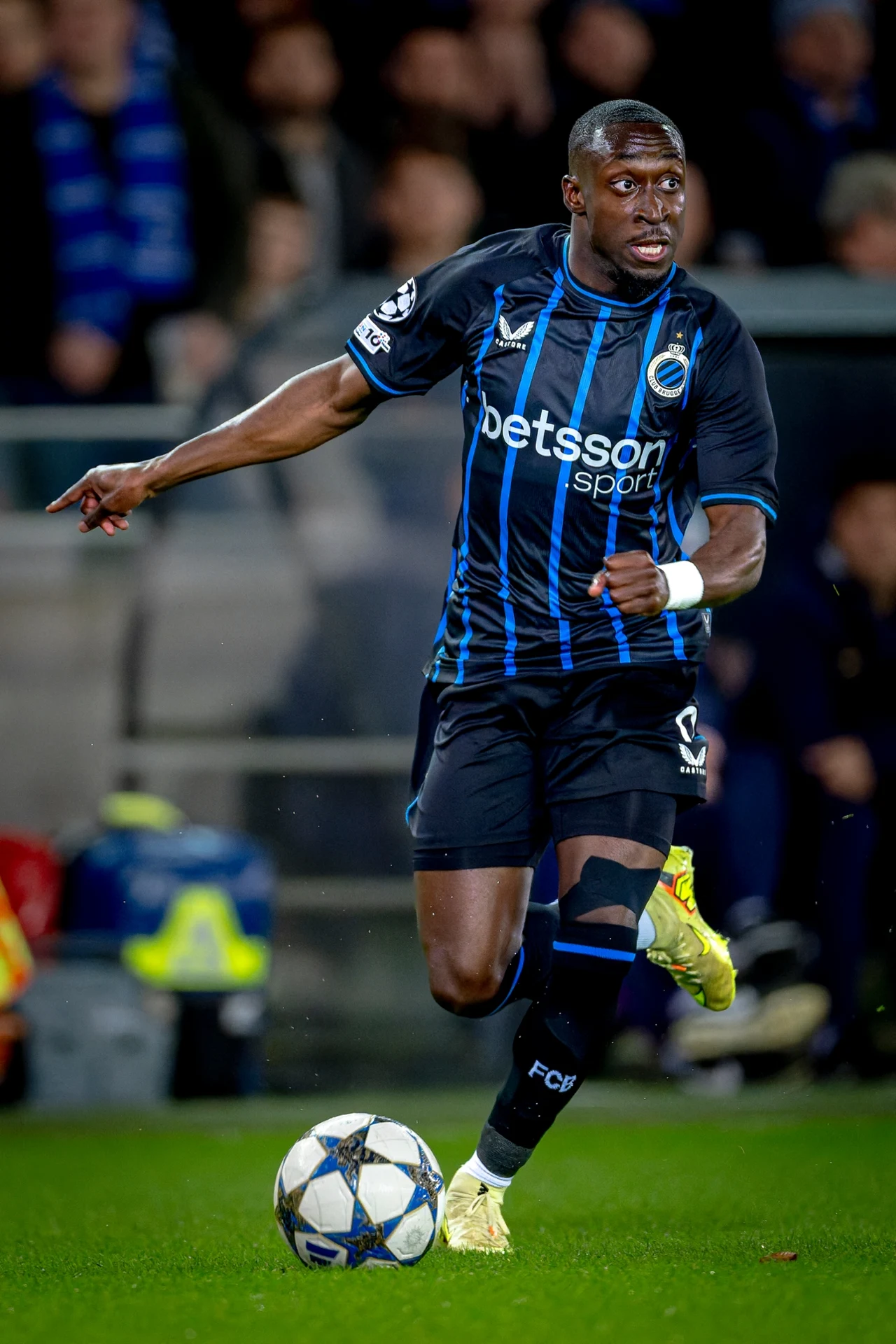 Piłkarz w czarno-niebieskim stroju drużyny Club Brugge biegnie po murawie z piłką podczas meczu, skupiony na akcji ofensywnej, w tle widoczni kibice oraz fragment trybuny stadionu.