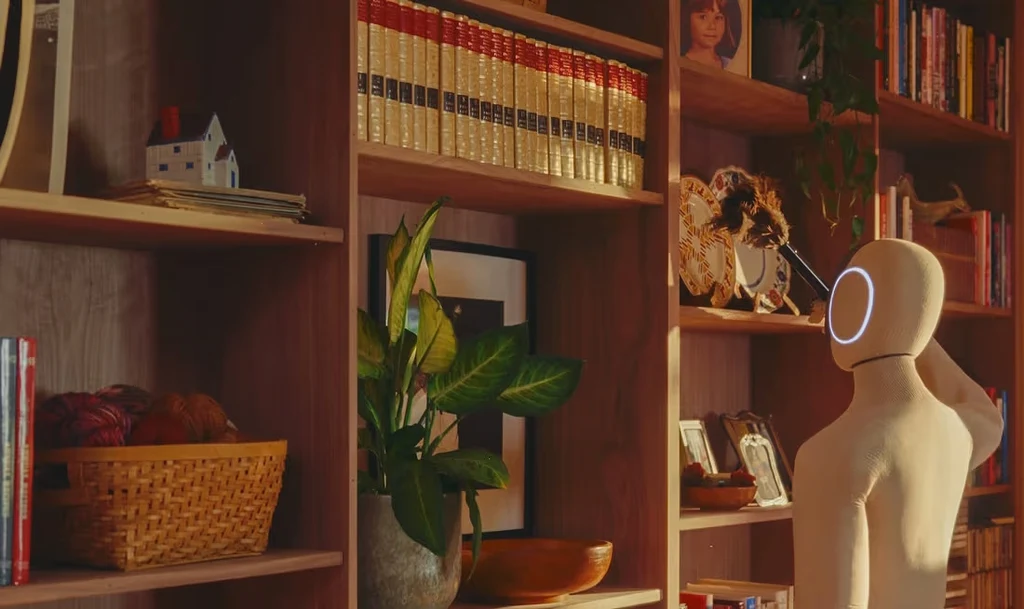 X1 NEO powstał, by ludzie mogli spędzać więcej czasu na aktywnościach, które naprawdę lubią Humanoidalny robot stoi przy drewnianym regale wypełnionym książkami, rośliną doniczkową oraz dekoracjami, spoglądając w stronę półek. W tle widoczne są różne przedmioty codziennego użytku, co buduje domową atmosferę.