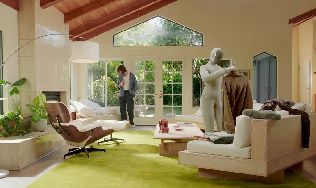 Według zapewnień producenta 1X NEO potrafi wiele, ale zawsze może nauczyć się więcej - wystarczy specjalny trenigng Jasny salon z dużymi oknami wychodzącymi na ogród, zielonym dywanem i drewnianym sufitem. Na sofie stoi biała figura ludzka o artystycznym charakterze, a w tle mężczyzna spoglądający na telefon. Wnętrze urządzone nowocześnie, z roślinami i dizajnerskim...