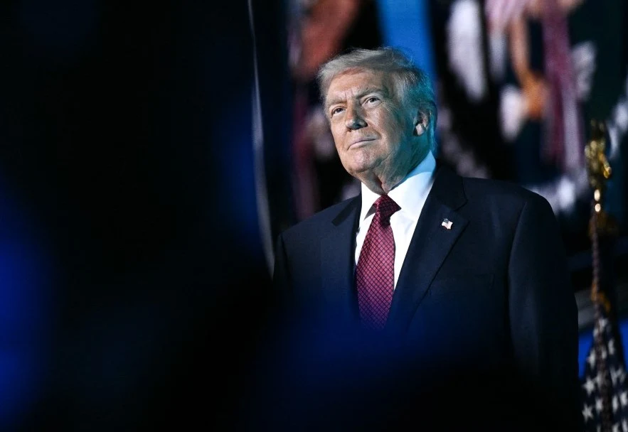 Mężczyzna w ciemnym garniturze, białej koszuli i czerwonym krawacie stoi w oświetlonym miejscu, patrząc lekko w górę, z poważnym wyrazem twarzy, na tle rozmytych flag i elementów sceny.