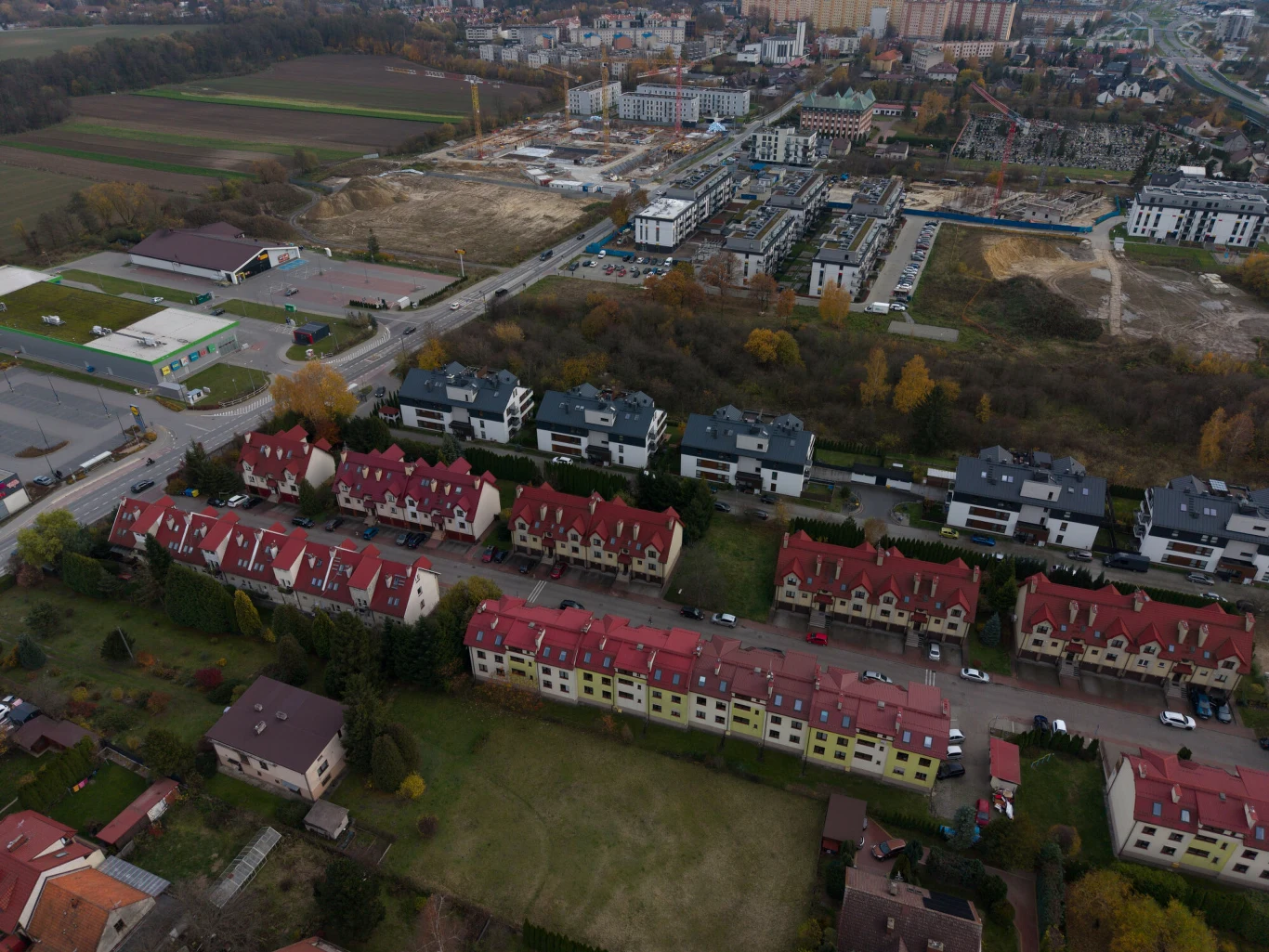 Samorządy zostawiają miliardy w budżetach (zdj. ilustracyjne) Zabudowa mieszkaniowa złożona z kilku rzędów jednolitych budynków z czerwonymi dachami, oddzielonych pasem zieleni od nowoczesnych osiedli apartamentowców. W tle rozległe tereny miejskie, place budowy i pola uprawne, jesienne drzewa oraz infrastruktura...