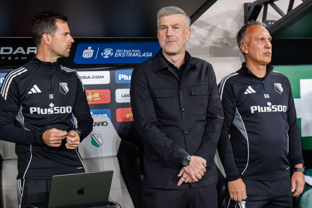 Edward Iordanescu (w środku) odszedł z Legii, a zastąpił go Inaki Astiz Trzech mężczyzn w sportowych strojach stojących przy ławce trenerskiej, dwóch z nich w bluzach z logo Legii Warszawa oraz sponsora, w tle tablice z logotypami partnerów Ekstraklasy.
