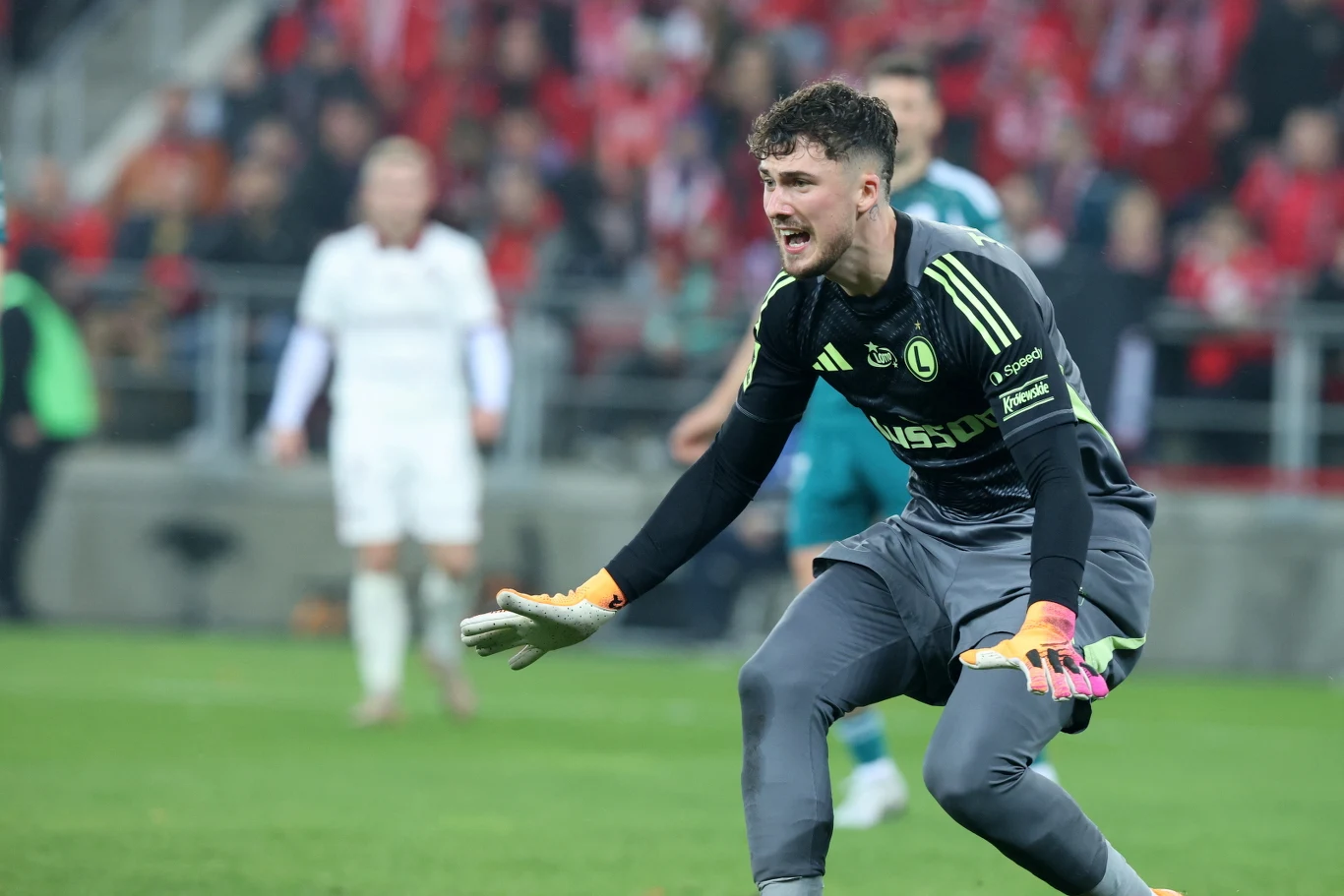 Kacper Tobiasz broni w Legii Warszawa Bramkarz w ciemnym stroju sportowym podczas meczu piłki nożnej, dynamicznie reaguje na sytuację na boisku, w tle rozmyte postacie innych zawodników i widzowie na trybunach.
