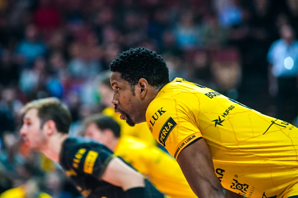 Zawodnik drużyny piłki ręcznej w żółtej koszulce z logo sponsorów obserwuje wydarzenia na boisku, skupiony na grze, z rozmytym tłem i innymi graczami w tle.