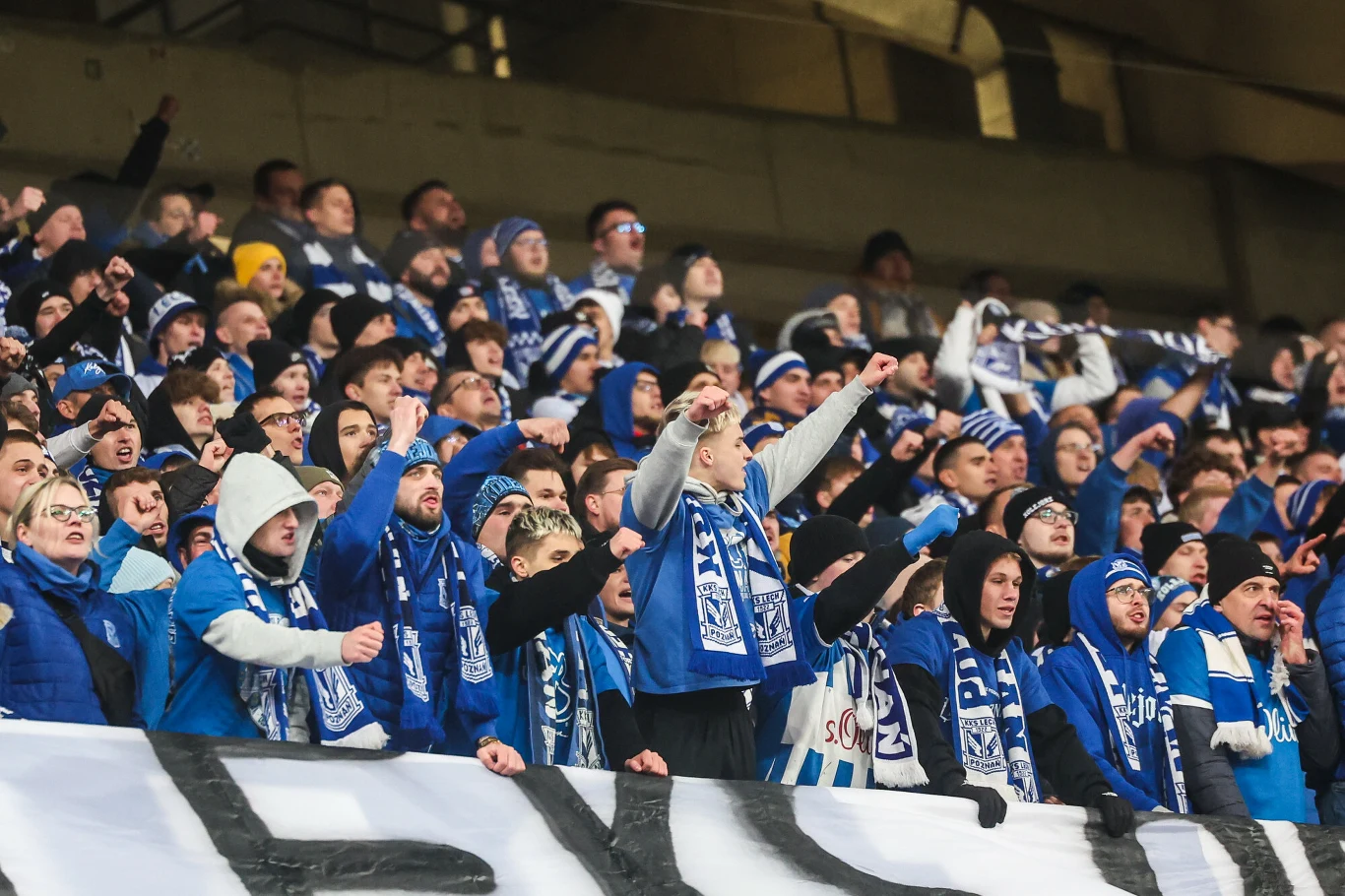 Kibice Lecha Poznań Tłum kibiców ubranych w niebiesko-białe szaliki i czapki energetycznie dopinguje swoją drużynę, z uniesionymi rękami, stojąc blisko siebie na trybunach stadionu sportowego.