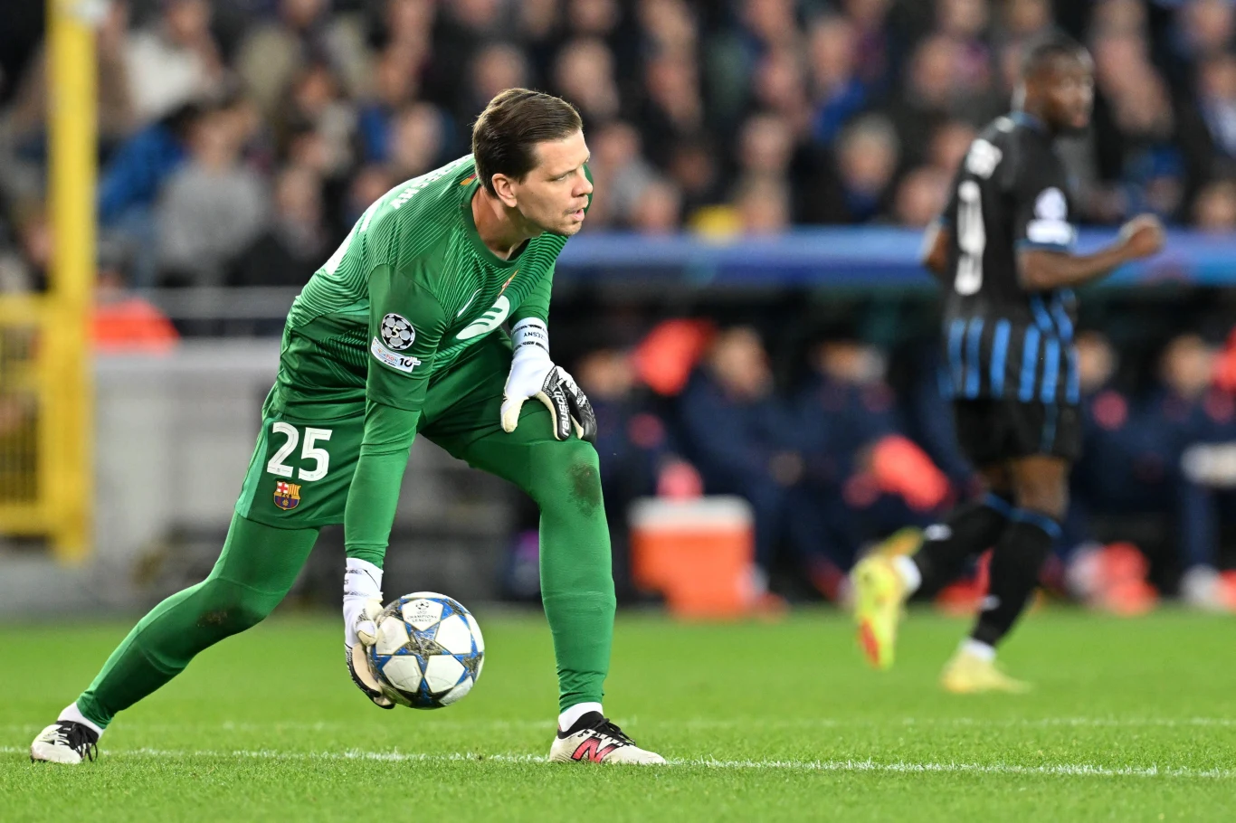 Bramkarz w zielonym stroju przygotowuje się do wybicia piłki na boisku piłkarskim podczas meczu, w tle widoczny inny piłkarz oraz publiczność na trybunach.