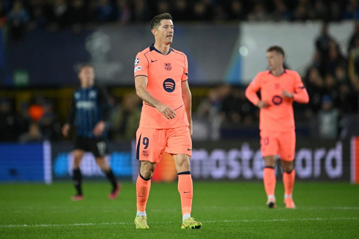 Piłkarz w różowym stroju drużyny FC Barcelona stoi na murawie boiska podczas meczu, w tle widoczni są inni zawodnicy oraz rozmyta publiczność.