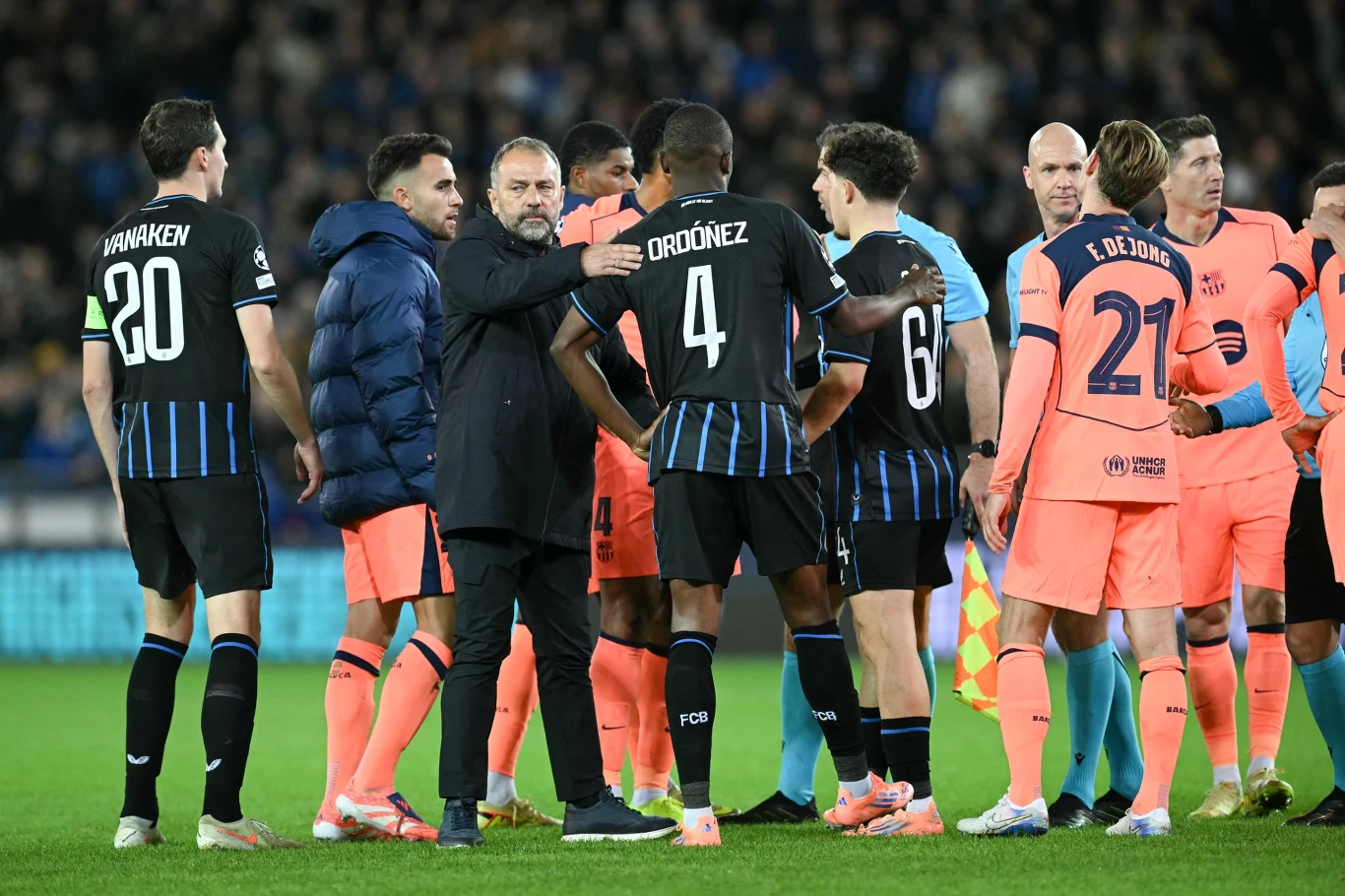 Piłkarze dwóch drużyn na murawie stadionu podczas przerwy lub po zakończeniu meczu, otoczeni przez sztab szkoleniowy, część zawodników ubrana w czarne stroje z niebieskimi paskami, a część w pomarańczowe zestawy, na twarzach sportowców skupienie i emocje.