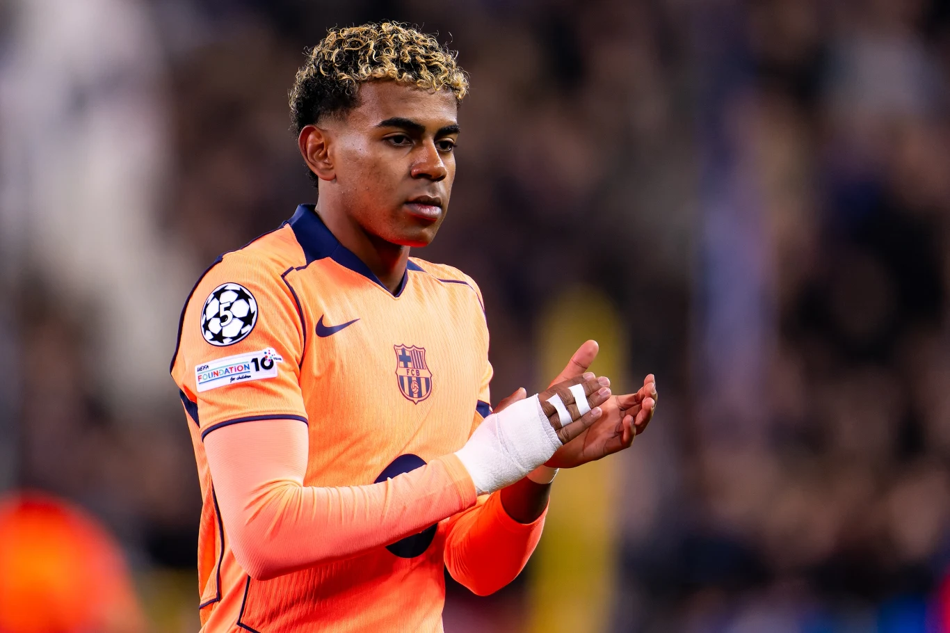 Piłkarz w pomarańczowej koszulce drużyny z emblematami Ligi Mistrzów UEFA, o krótkich kręconych włosach, z bandażem na dłoni, stoi na boisku, skupiony, w tle rozmyte postacie kibiców.