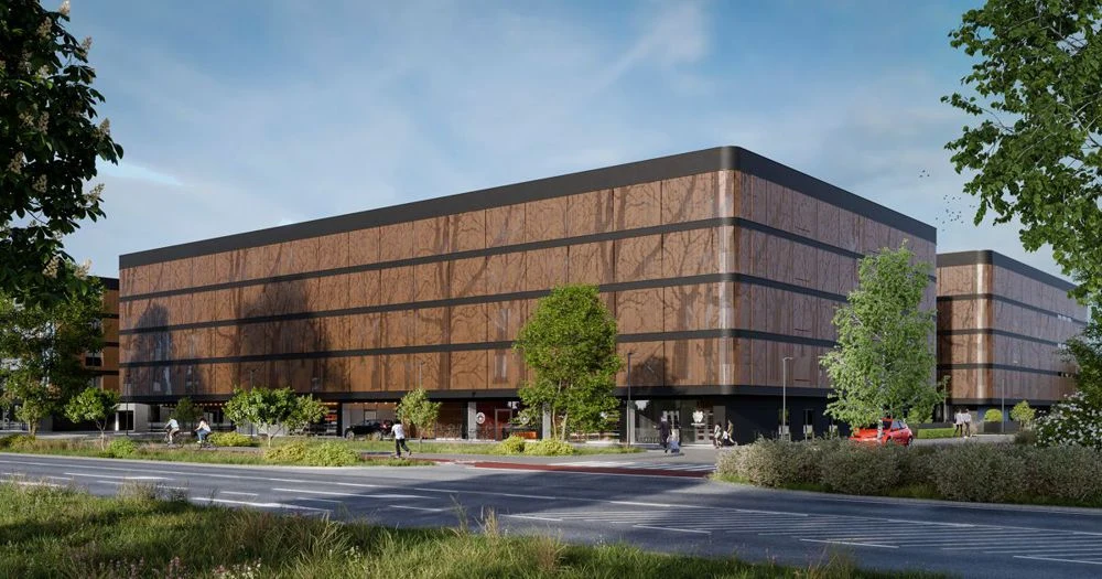 Błonia Residence Nowoczesny, prostokątny budynek z przeszkloną elewacją w odcieniach brązu, otoczony zielenią i drzewami, przy szerokiej ulicy. Na chodniku widać kilku przechodniów, a przed budynkiem zaparkowane są samochody.