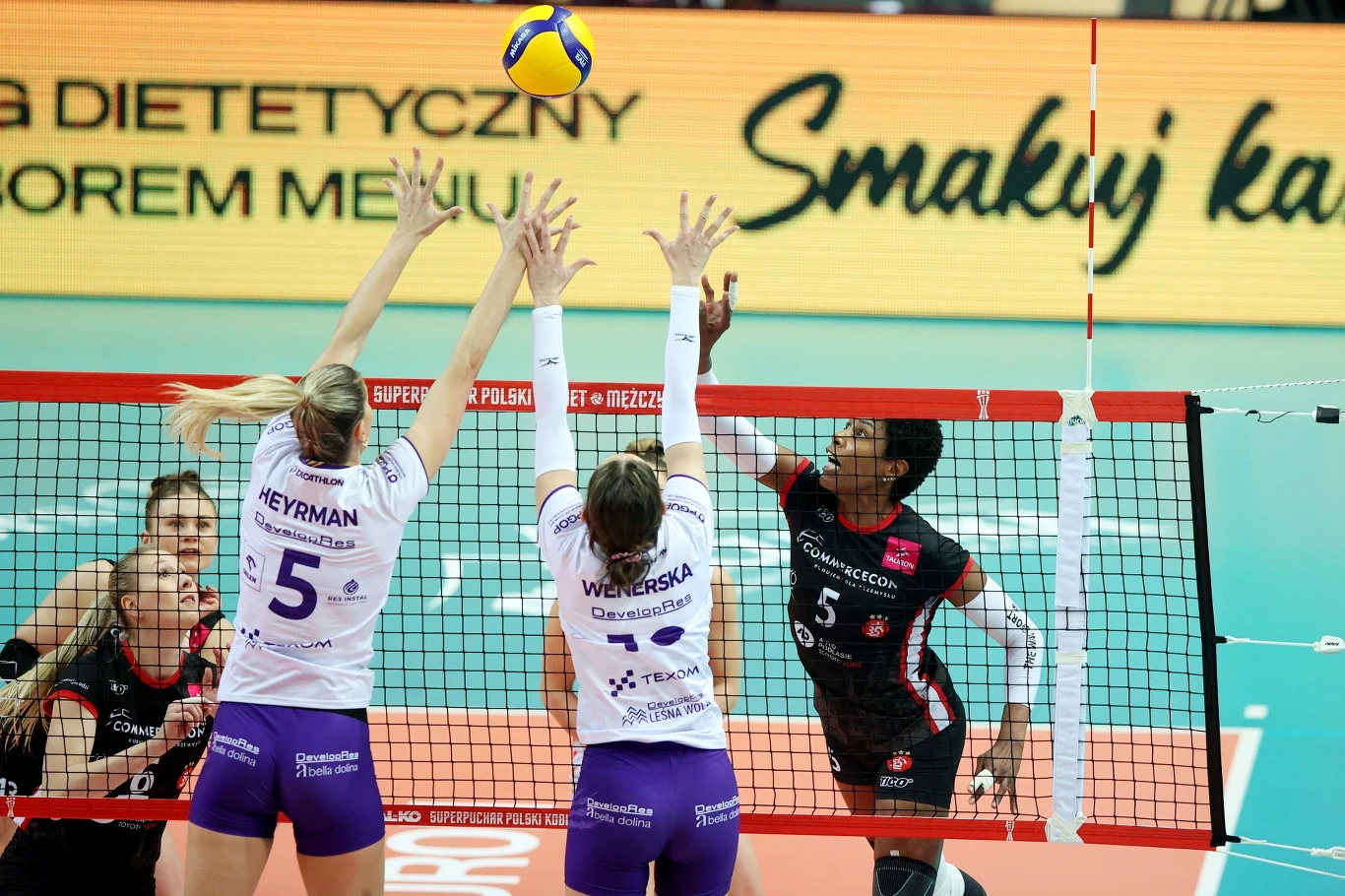 Laura Heyrman (z lewej) w bloku przy ataku Regiane Bidias z ŁKS-u Commercecon Łódź Siatkarki rywalizują przy siatce, dwie zawodniczki drużyny w białych koszulkach próbują zablokować atakującego zawodnika drużyny przeciwnej uderzającego piłkę, dynamiczna akcja na tle parkietu sportowego.