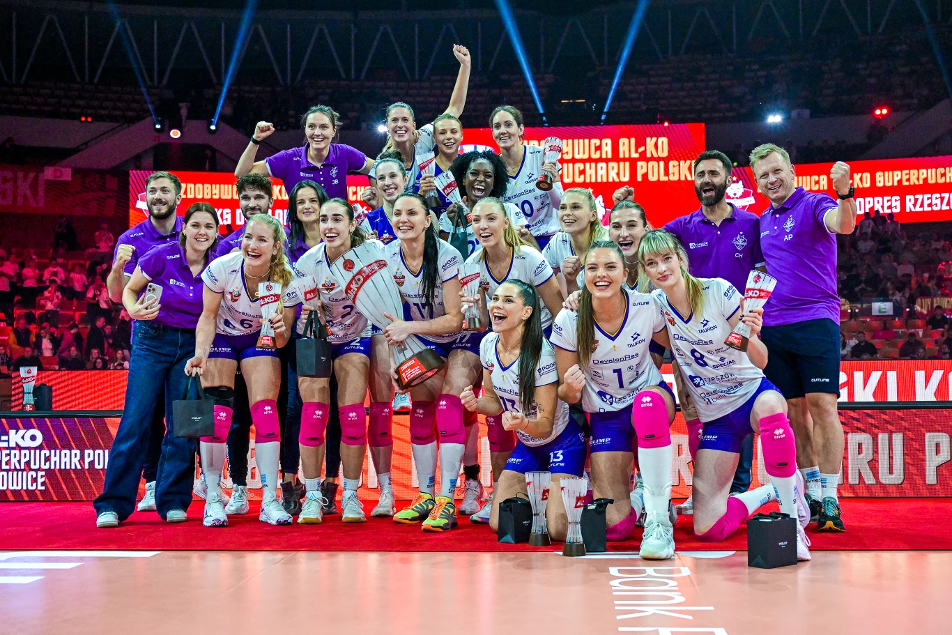 Siatkarki DevelopResu Rzeszów po raz trzeci w historii sięgnęły po AL-KO Superpuchar Polski Drużyna siatkarek w białych i fioletowych strojach świętuje zwycięstwo na hali sportowej, w tle widoczne czerwone banery i napisy z nazwą rozgrywek, zawodniczki uśmiechnięte, niektóre z uniesionymi rękami.