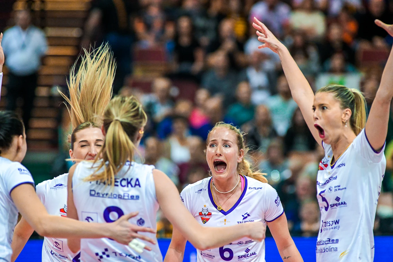 Zawodniczki drużyny siatkarskiej w białych strojach gromadzą się na boisku, wyrażając radość i emocje po udanej akcji, otoczone trybunami z widownią w tle.