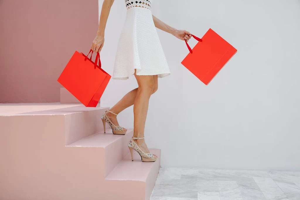 Zadbaj o bezpieczeństwo zakupów online Kobieta w białej sukience i eleganckich butach schodzi po jasnoróżowych schodach, trzymając w dłoniach dwie czerwone torby na zakupy.