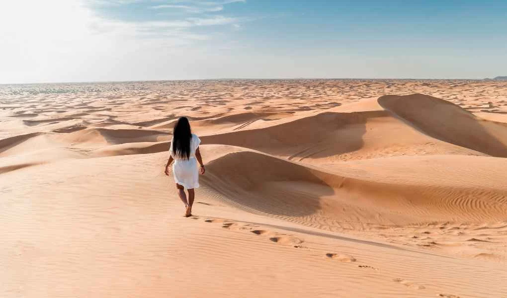 Kobieta w białej sukience idzie po pustyni, zostawiając ślady na piasku, wokół rozciągają się piaszczyste wydmy oświetlone jasnym światłem słonecznym, a na horyzoncie widać czyste, niebieskie niebo.