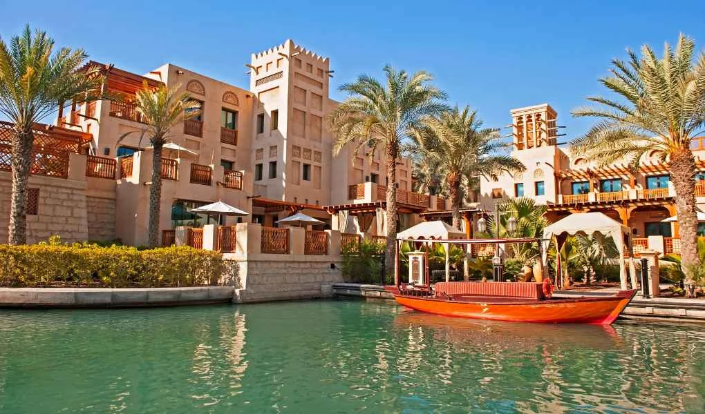 Luksusowy hotel w stylu arabskim z charakterystycznymi wieżami, otoczony palmami, przy kanale wodnym z tradycyjną łodzią na wodzie, wszystko w otoczeniu egzotycznej zieleni i słonecznej pogody.