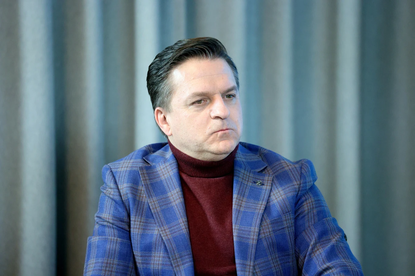 Bogdan Rymanowski Mężczyzna w średnim wieku w niebieskiej marynarce w kratę i bordowym golfie, siedzący na tle pionowych, stonowanych zasłon, z poważnym wyrazem twarzy.