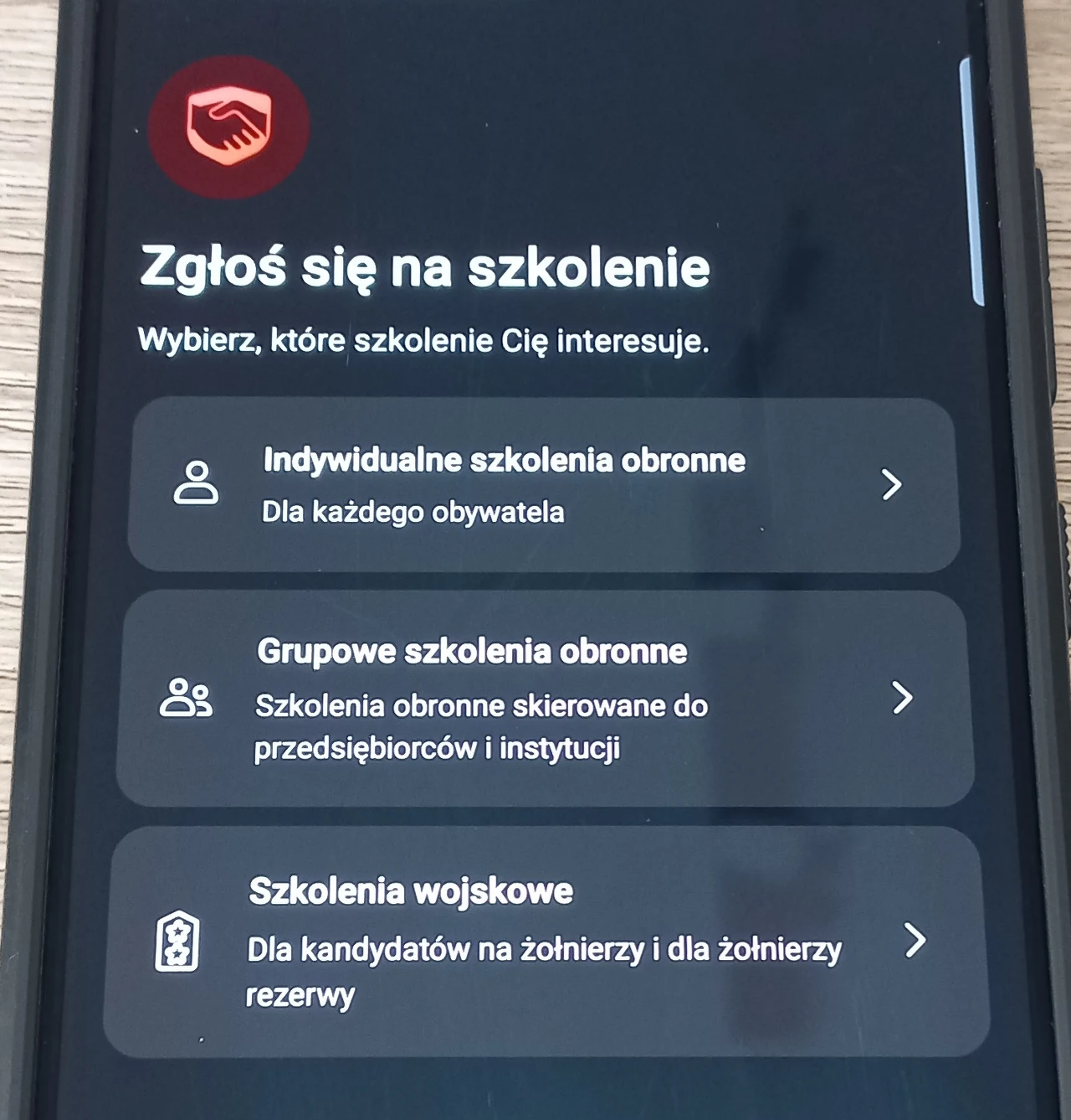 Panel zgłoszenia w mObywatelu. Ekran telefonu z aplikacją dotyczącą szkoleń obronnych, zawierającą trzy opcje do wyboru: indywidualne szkolenia obronne dla każdego obywatela, grupowe szkolenia obronne dla przedsiębiorców i instytucji oraz szkolenia wojskowe dla kandydatów na żołnier...