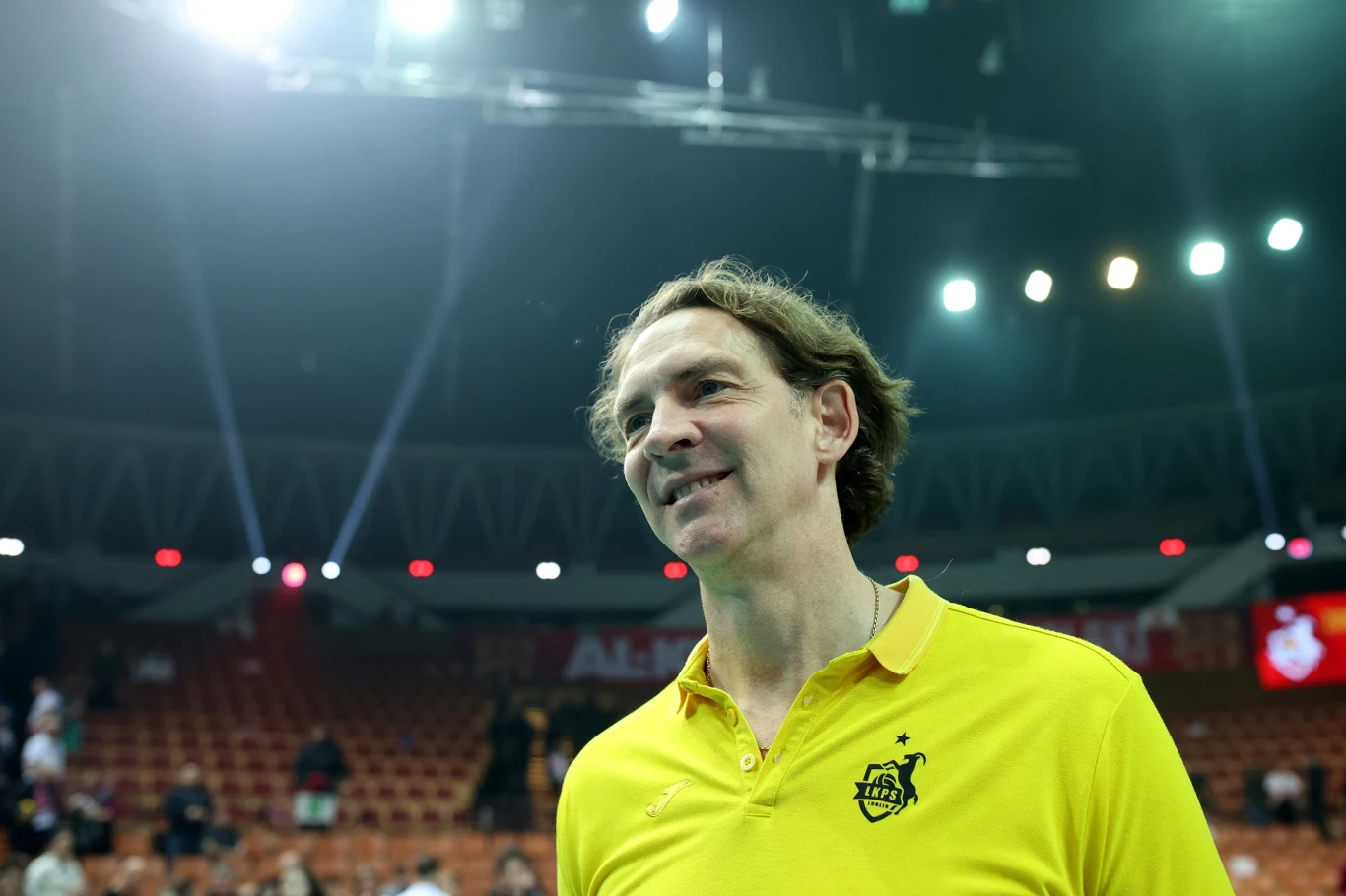 Mężczyzna w żółtej koszulce z logo sportowym stoi na hali sportowej, w tle widoczne rzędy pustych krzeseł oraz kilka osób. Oświetlenie sufitowe tworzy jasną atmosferę wnętrza.