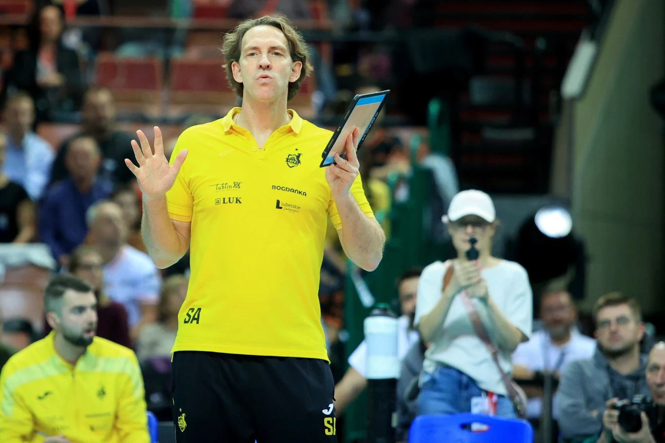 Trener w żółtej koszulce z tabletem w ręku podczas meczu siatkarskiego, w tle siedzący kibice oraz stojąca fotografka z aparatem.