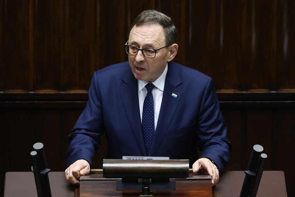 Lider mniejszości niemieckiej Ryszard Galla Mężczyzna w garniturze przemawia przy mównicy, w tle widoczny drewniany panel. Osoba ma na sobie okulary oraz krawat w kropki.