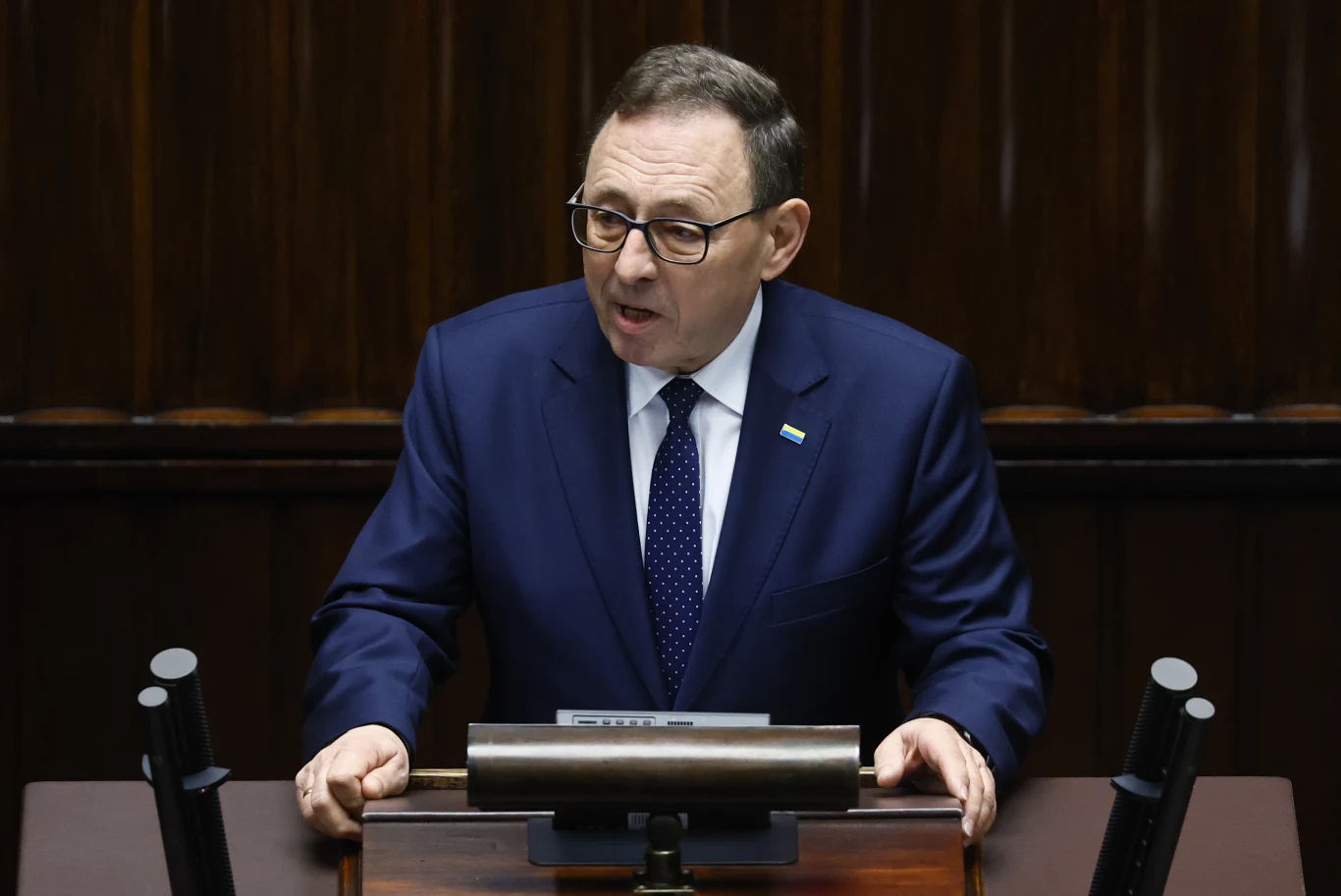 Lider mniejszości niemieckiej Ryszard Galla Mężczyzna w garniturze przemawia przy mównicy, w tle widoczny drewniany panel. Osoba ma na sobie okulary oraz krawat w kropki.