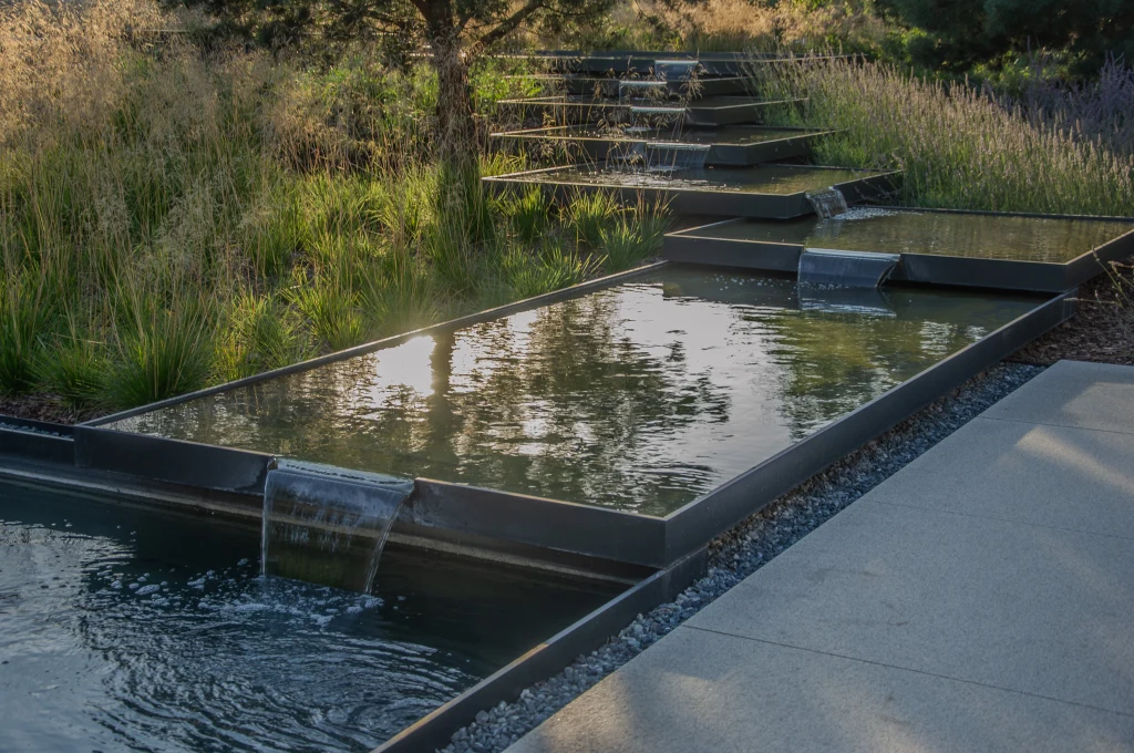 "Leśna wyspa" Szereg kaskadowo ułożonych prostokątnych basenów wodnych z delikatnie spływającą wodą, otoczonych bujną zielenią i trawami ozdobnymi w promieniach zachodzącego słońca.