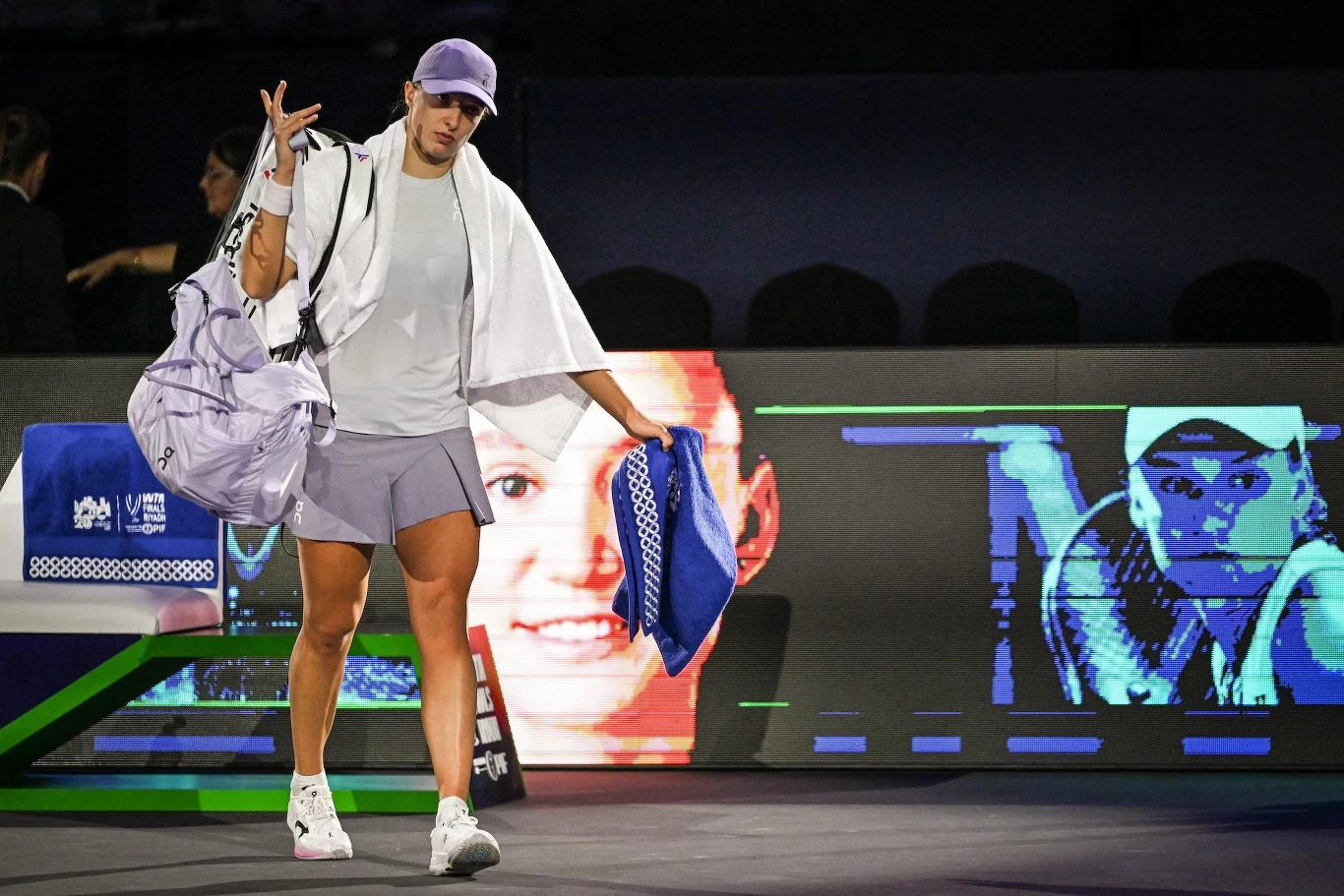 Tenisistka w stroju sportowym, z ręcznikiem na ramieniu i torbą sportową, schodzi z kortu na tle dużego ekranu wyświetlającego jej wizerunek. Oświetlenie skupione na zawodniczce, atmosfera turniejowa.